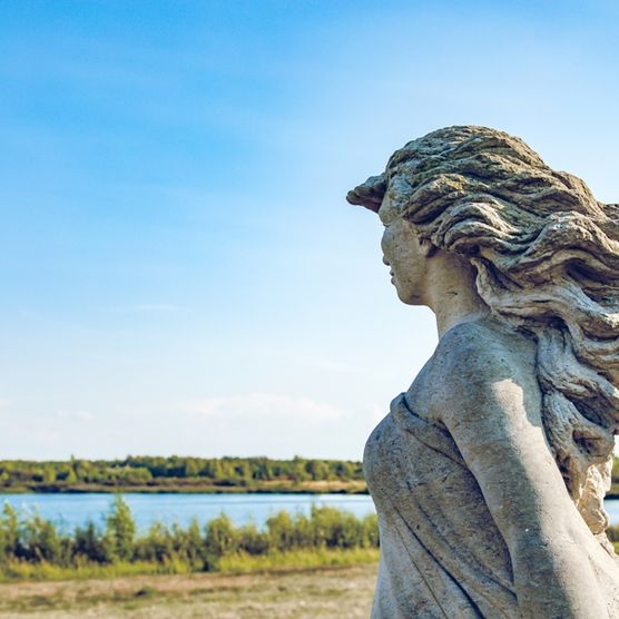 Das Bild zeigt eine Nah-Ansicht in welcher über die Schulter der Penelope-Statue Richtung See fotografiert wird.