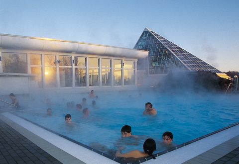 Das Außenbecken eines Schwimmbades ist voller Badegäste, der heiße Dampf steigt auf und im Hintergrund ist Abendhimmel zu erkennen.