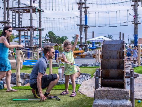 Das Bild zeigt eine junge Familie beim Mini-Golf Spielen. Der Vater hilft seiner Tochter beim Zielen. 