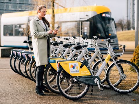 Eine Frau hält ihr Handy in der Hand und steht neben vielen nextbike-Leihfahrrädern. Im Hintergrund fährt eine Tram.