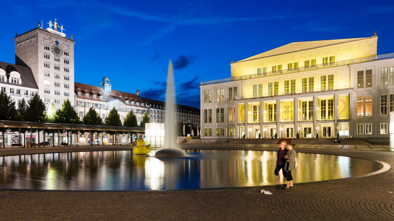 Ein Bild der Oper Leipzig bei Dämmerung auf dem Augustusplatz. Das Gebäude ist hell erleuchtet und reflektiert im Brunnen vor dem Opernhaus, während der Himmel in tiefem Blau erstrahlt.