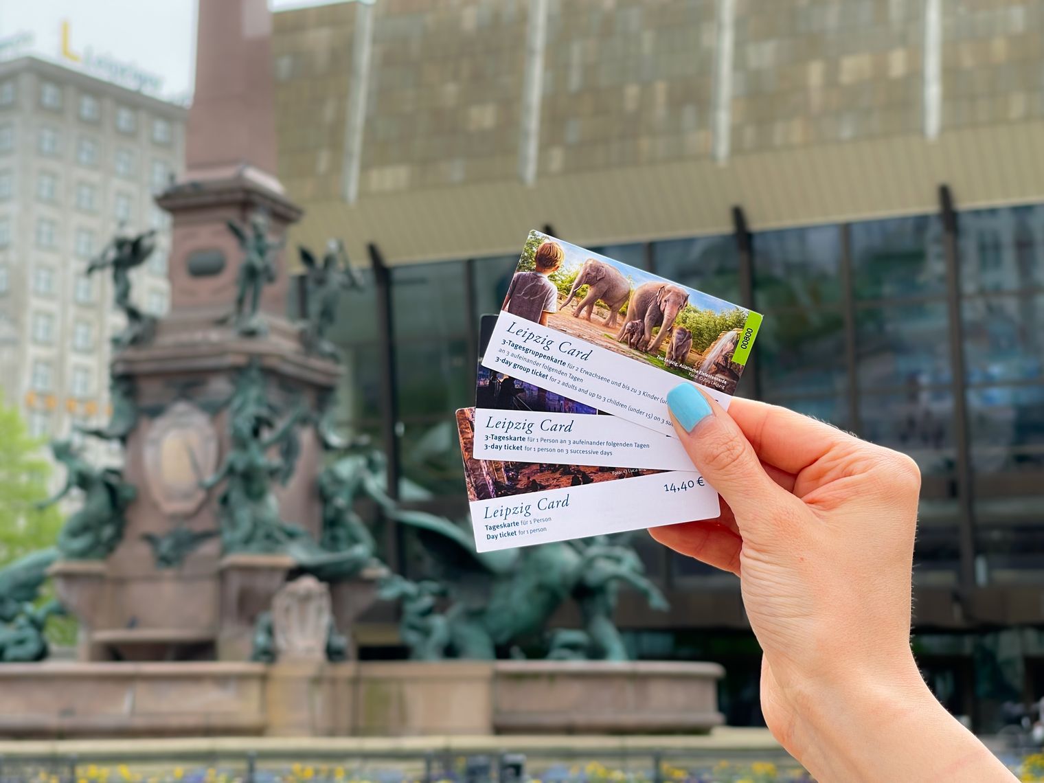 Eine Hand hält alle erhältlichen LEIPZIG CARDS vor dem Mendebrunnen auf dem Augustusplatz in die Höhe