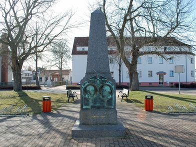 Denkmal für Martin Luther und Katharina von Bora in Neukieritzsch, zu sehen ist eine Stele mit den Porträts der Beiden auf dem Marktplatz mit Parkbänken und Fahrradständern © LTM, Jasmin Rhein