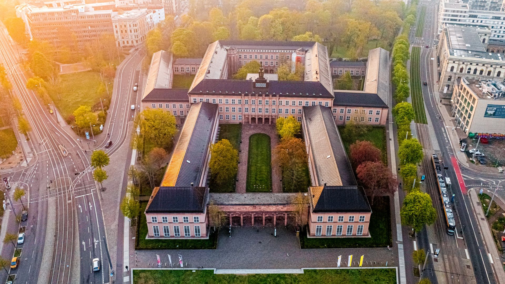 Die Luftaufnahme zeigt den Gebäudekomplex der Museen im GRASSI in rötlichen Tönen und umgeben von grünen Bäumen von oben