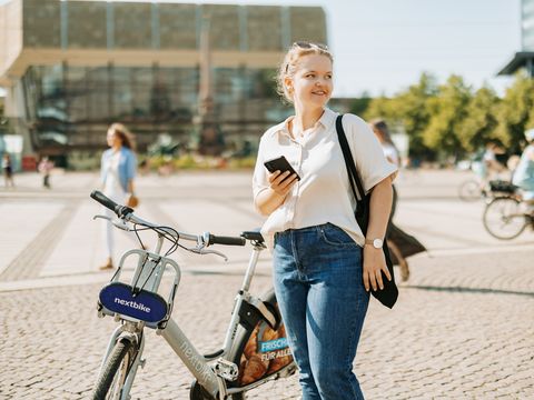 Eine junge Frau steht auf dem Augustusplatz in Leipzig. Sie hält lächelnd ihr Handy in der Hand, neben ihr steht ein Nextbike-Leihfahrrad. 