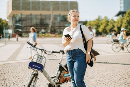 Eine junge Frau steht auf dem Augustusplatz in Leipzig. Sie hält lächelnd ihr Handy in der Hand, neben ihr steht ein Nextbike-Leihfahrrad. 