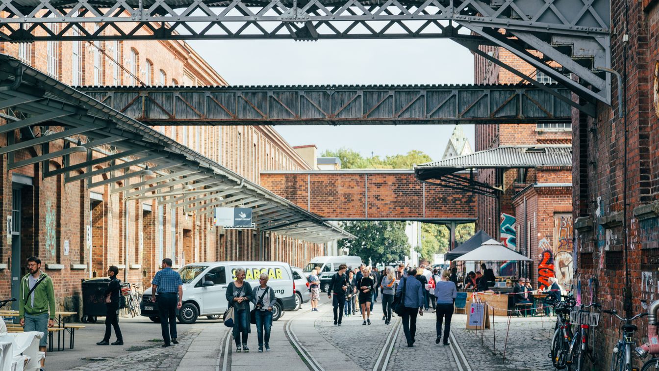 Mehrere Menschen laufen beim Spinnereirundgang über das Gelände der ehemaligen Leipziger Baumwollspinnerei in Leipzig Plagwitz. Links und rechts sieht man die früheren Industriegebäude, dazwischen befindet sich gepflasterter Weg mit Bahnschienen