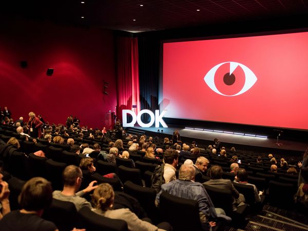 Eröffnung DOK Leipzig 2019, zu sehen ist ein vollbesetzter Leipziger Kinosaal sowie eine rote Leinwand mit dem Bild eines Auges, davor steht in großen Buchstaben "DOK" © Susann Jehnichen