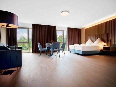 Ein modern gestaltetes Komfortzimmer mit Bett, Sitzgarnitur, Schreibtisch und langen braunen Vorhängen im Hostel Platsch in Oschatz.