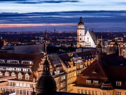 Ein Bild der Thomaskirche in Leipzig bei Dämmerung. Die Kirche ist beleuchtet und hebt sich eindrucksvoll gegen den dunklen Himmel ab.