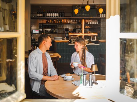 Durch das geöffnete Fenster fällt der Blick von außen in den gemütlichen Gastraum, wo zwei junge Menschen am Tisch sitzend Kaffee trinken.
