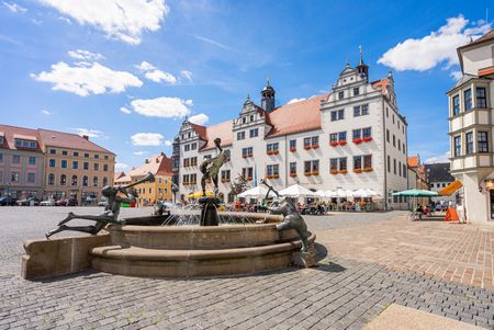 Das Bild zeigt Dem Marktplatz von Torgau. In der Mitte des Platzes steht ein großer Brunnen, rechts dahinter im Bild sieht man die weiße Fassade des städtischen Rathauses.