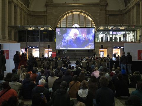 In der Eingangshalle des Leipziger Hauptbahnhofs ist eine große Leinwand aufgebaut vor der Menschen auf Stühlen einen Film schauen. Hinter ihnen sitzen weitere Menschen auf dem Boden und sehen sich ebenfalls den Film an. Das Licht ist gedimmt.