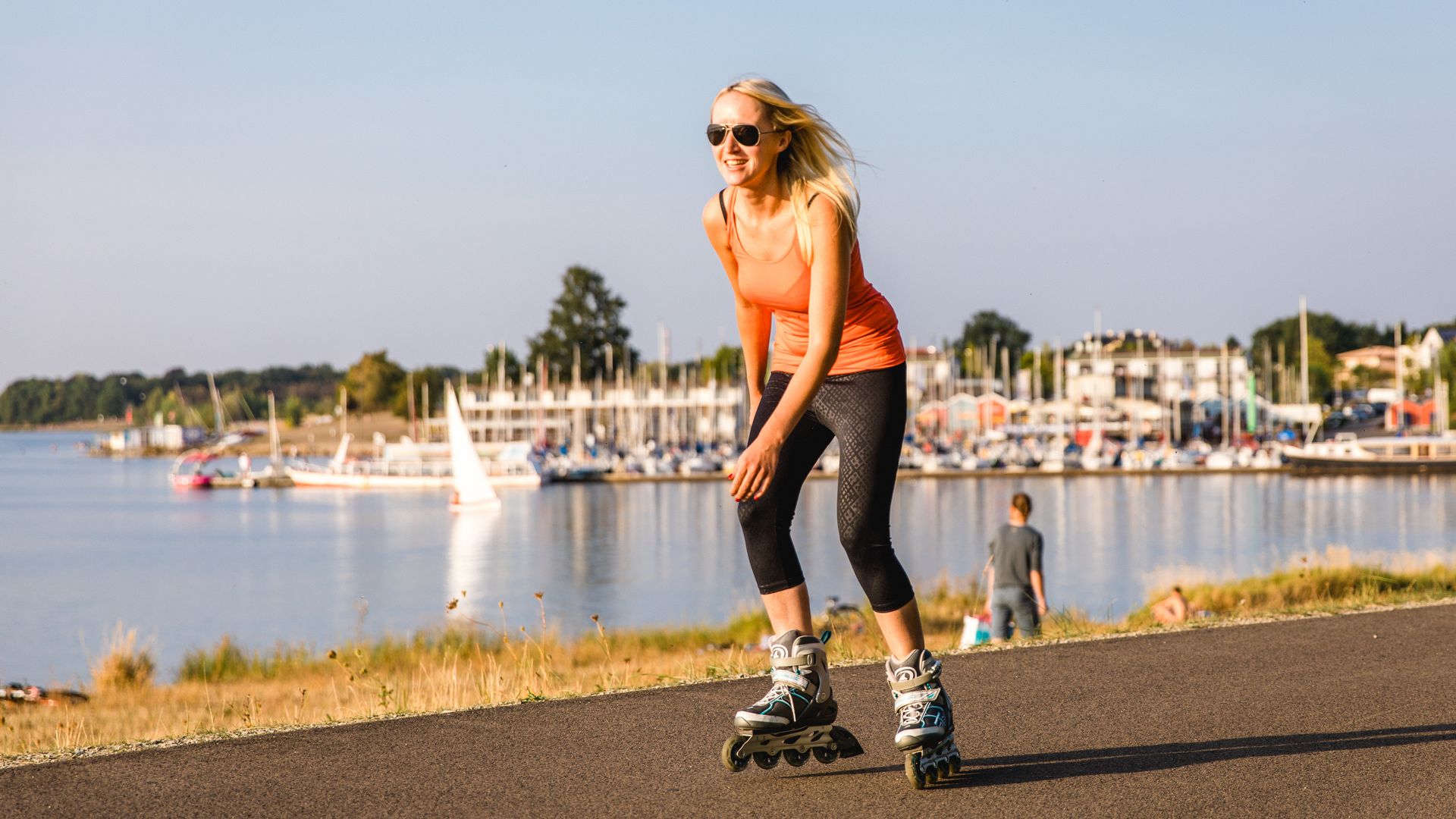 Eine junge Frau fährt auf dem asphaltierten Rundweg am Südufer des Cospudener Sees Inlineskates, im Hintergrund sieht man die Marina des Hafen Zöbigker, auch Pier 1 genannt
