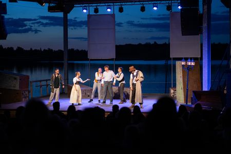 Das Bild zeigt eine Gruppe von sechs Schauspielern auf der Seebühne des Biedermeierstrands, im Hintergrund das dunkle Wasser des Schladitzer Sees bei Abenddämmerung.