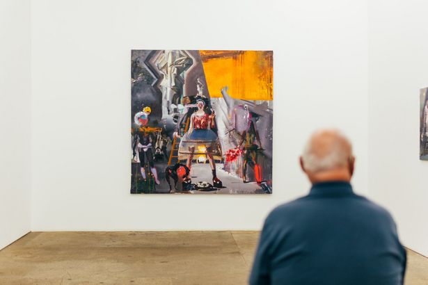 An elderly man sits in front of an abstract painting of a woman in a gallery in the Spinnerei