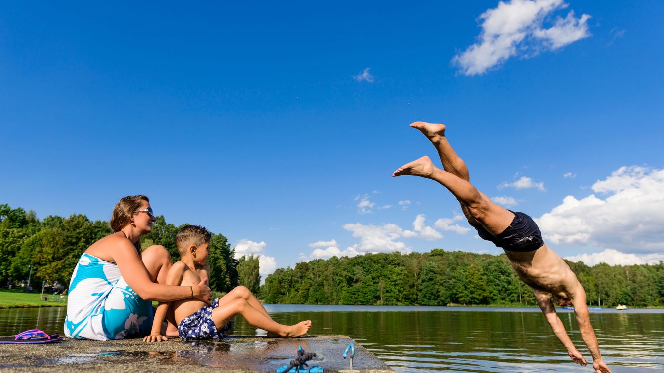 Eine Familie mit Kindern sitzt entspannt an einem naturbelassenen See und genießt den Familienausflug an einem Sommertag, während der Vater kopfüber ins Wasser springt. 