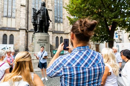 Ein junger Mann und eine junge Frau stehen während einer Stadtführung am Bach-Denkmal an der Thomaskirche Leipzig und hören einem Gästeführer zu, der Mann macht ein Foto mit seinem Smartphone. 