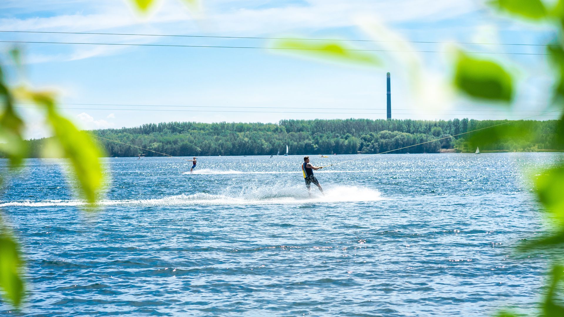 Eingerahmt von grünen Baumblättern zeigt das Bild den Blick auf den Kulkwitzer See in Leipzig, im Mittelpunkt des Bildes sieht man einen Mann auf einem Wakeboard, der sich von der Wasserskiwinde über das Wasser ziehen lässt