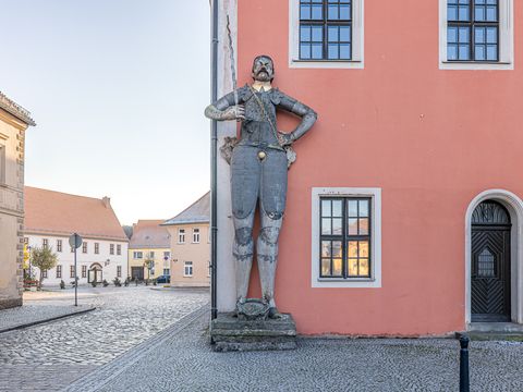 Das Bild zeigt eine Roldandfigur an der Ecke des rot-verputzten Rathauses in Belgern, im linken Bereich sieht man die historische ALtstadt des Ortes.