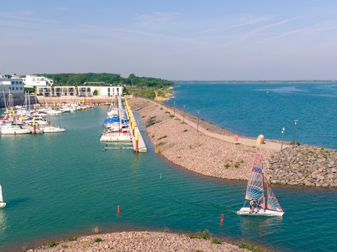 Das Bild zeigt eine malerische Hafenszene an einem sonnigen Tag. Im Vordergrund ist ein kleiner Segelboot mit einem weißen Segel zu sehen, das auf dem ruhigen, türkisfarbenen Wasser segelt. Rechts davon befindet sich ein weiteres Segelboot mit einem bunten Segel in Blau und Orange. Der Hafen ist von einer langen Mole aus Steinen umgeben, die ins Wasser ragt und den Hafenbereich vom offenen Gewässer trennt. Auf der Mole verläuft ein gepflasterter Weg mit Laternen, der entlang des Wassers führt. Im Hintergrund sind mehrere größere Boote und Yachten vertäut, die an gelben Pfählen festgemacht sind. Hinter den Booten stehen moderne Gebäude mit flachen Dächern, die wahrscheinlich Wohn- oder Geschäftsgebäude sind. Weiter im Hintergrund erstreckt sich eine grüne Landschaft mit Bäumen und Hügeln unter einem klaren blauen Himmel. Die gesamte Szene strahlt Ruhe und Gelassenheit aus, typisch für einen entspannten Tag am Wasser.