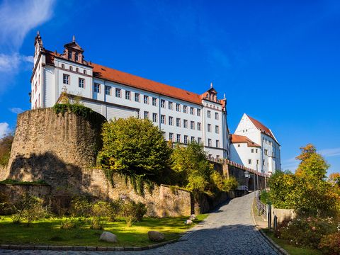 Blick auf das Schloss Colditz mit weißen Fassaden und roten Dächern, umgeben von einer alten Steinmauer und einem gepflasterten Weg.