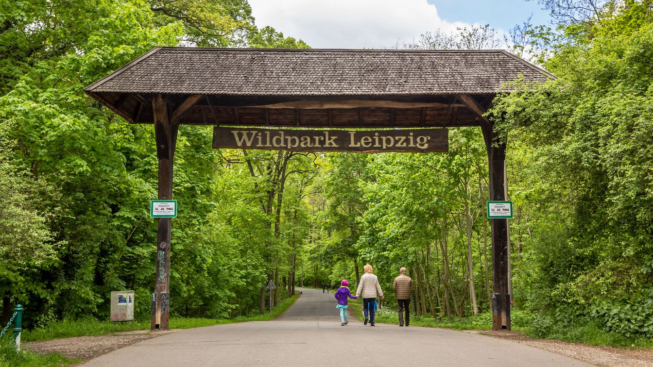Ein großer Holzbogen markiert den Eingang zum Wildpark Leipzig, umgeben von dichtem Grün und drei Personen sind auf dem Weg in den Park zu sehen.