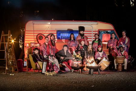 Eine zwölfköpfige Musikgruppe mit diversen Instrumenten und Wohnzimmer-Möbeln steht vor einem orange-rot erleuchteten Wohnwagen, es brennt eine Feuerschale