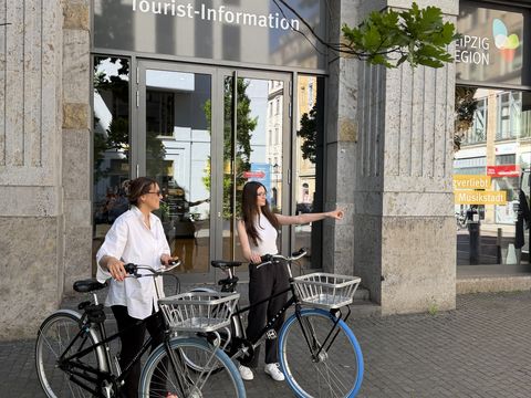 Zwei junge Frauen mit Swapfiets-Fahrrädern vor dem Eingang einer Tourist-Information Leipzig, eine zeigt in eine Richtung.