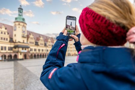 Ein Kind mit einem Smartphone fotografiert auf dem Marktplatz in Leipzig das Alte Rathaus.