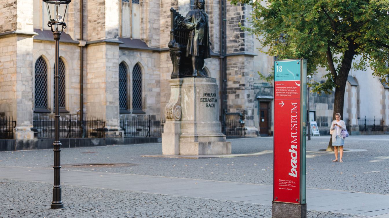 Das Bild zeigt einen Teil der Leipziger Notenspur mit Bachdenkmal, Thomaskirche im Hintergrund und einer Stele mit Informationen zum Bach-Museum.