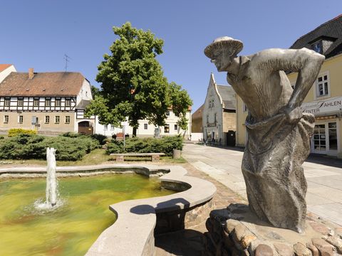Das Bild zeigt eine Seitenansicht der Bronzeskulptur eines Sackhuppers (Sackhüpfers) mit dem Dahlener Markt im Hintergrund. 