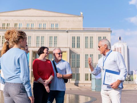 Ein Gästeführer von Leipzig Erleben spricht zu einer Gruppe vor der Oper Leipzig auf dem Augustusplatz