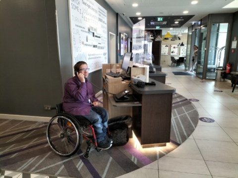 Ein Mann sitzt im Rollstuhl an einem niedrig gestellten Rezeptionstresen. Er telefoniert.