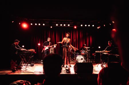 Auf einer Bühne steht eine Sängerin mit geschlossenen Augen und singt. Im Hintergrund steht eine Band bestehend aus 4 Personen. Von links nach Rechts: Ein Keyboarder, ein Gittarrist, ein Schlagzeuger und eine Musikerin am Synthesizer.