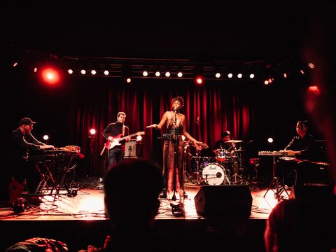 A singer is standing on a stage with her eyes closed and singing. In the background is a band consisting of 4 people. From left to right: a keyboard player, a guitarist, a drummer and a musician on the synthesizer.