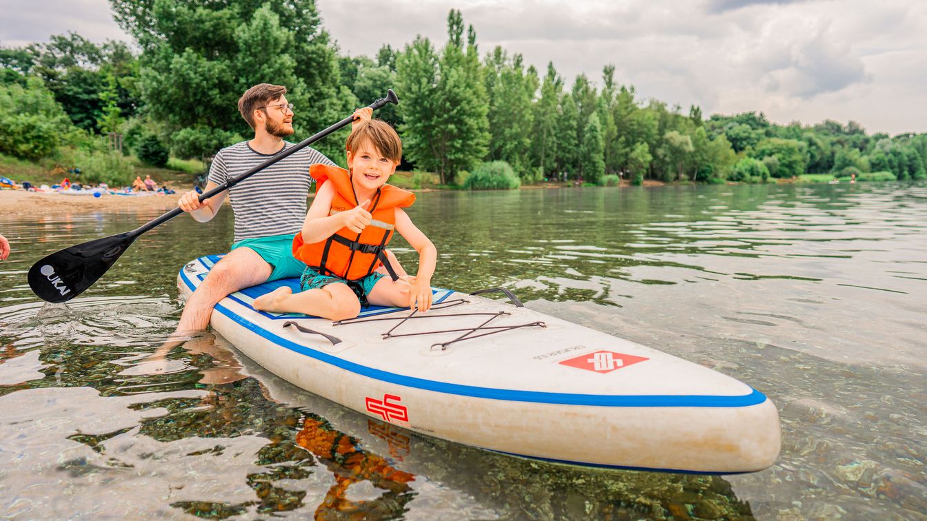 Ein Mann und ein Kind sitzen auf einem Stand-Up-Paddling-Board und fahren auf dem Cospudener See hinaus.