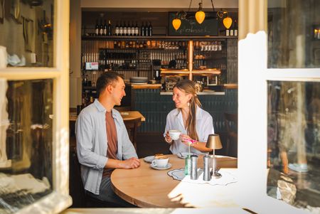 Blick von außen durch ein geöffnetes Fenster in das Restaurant und Café „Zum Arabischen Coffe Baum“, in dem ein junges Paar an einem Tisch direkt am Fenster sitzt und sich unterhält, während es seinen Kaffee genießt.
