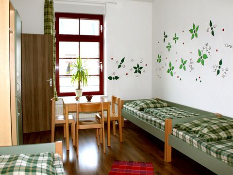 Blick auf ein helles Hostelzimmer mit zwei Einzelbetten, einem Tisch, Stühlen und Wanddekorationen.