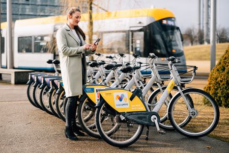Eine Person steht vor einer Reihe nextbikes und ist im Begriff, sich eines über das Handy auszuleihen. ImHintergrund fährt eine Straßenbahn.