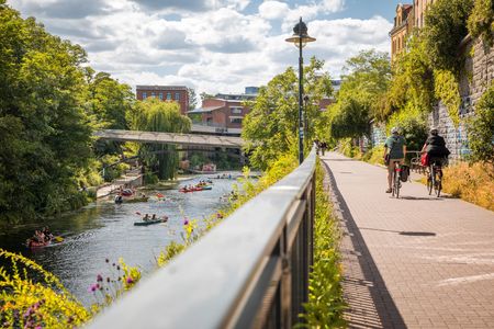 Das Bild zeigt den Karl-Heine-Kanal mit Kanu-Fahrern und den angrenzenden Radweg mit zwei Fahrradfahrern.