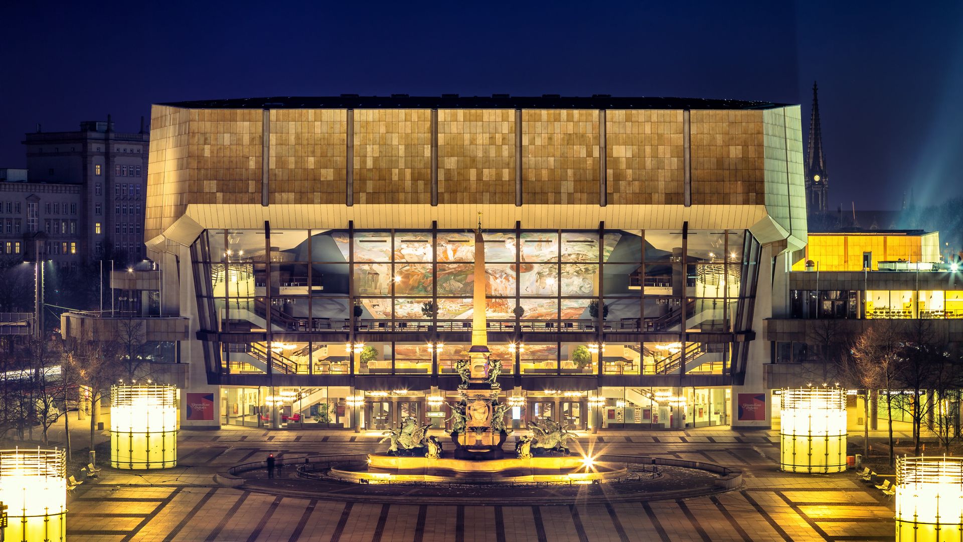 Pohled na osvětlenou kašnu Mendebrunnen a osvětlený lipský Gewandhaus, ve kterém hraje slavný Orchestr Gewandhausu a který je skutečnou pamětihodností města hudby Lipska, na náměstí Augustusplatz ve večerních hodinách, kulturní zařízení