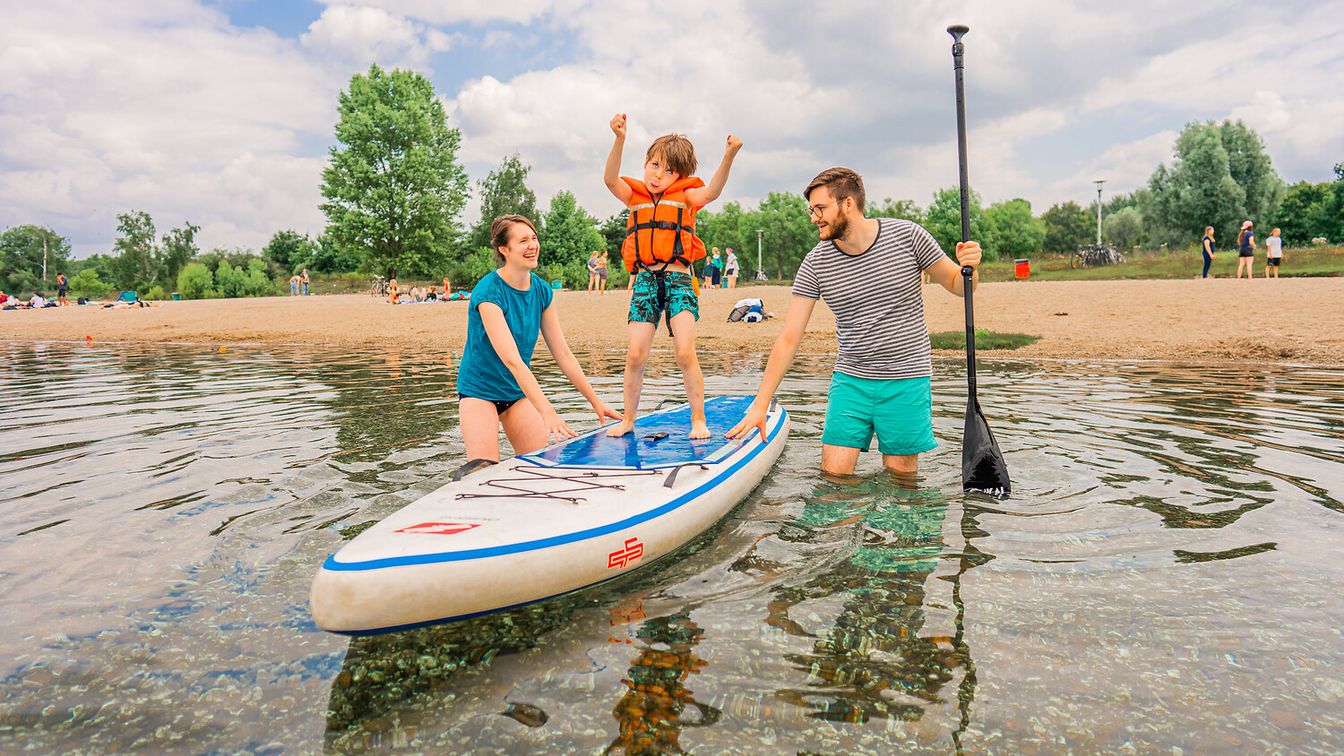 Ein Kind albert auf einem SUP auf dem Cospudener See herum. Die Eltern stehen mit im Wasser.