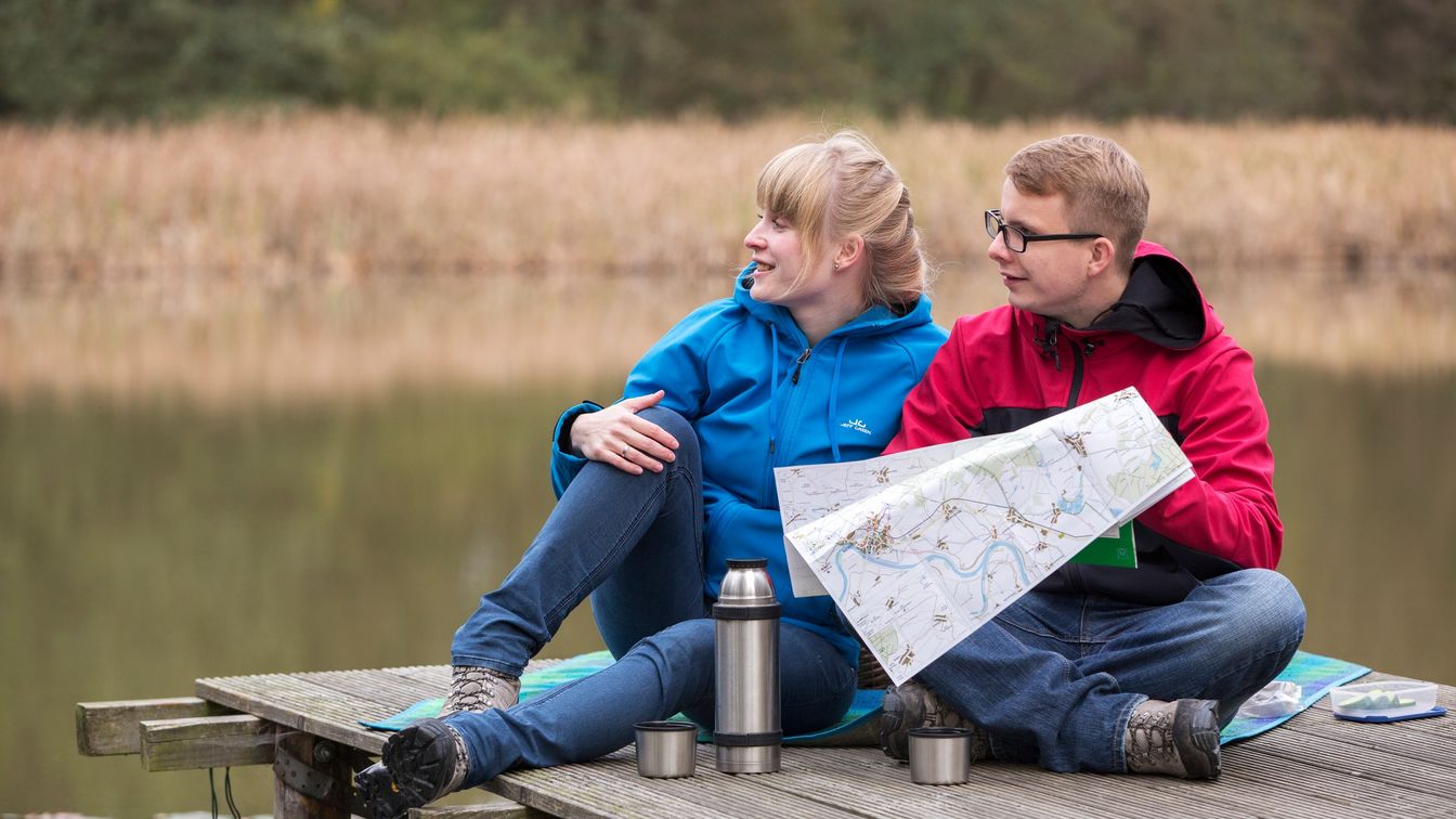 Das Bild zeigt zwei Wanderer mit Karte, die auf einem Steg über einem Teich in der Dahlener Heide sitzen.