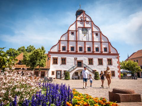 Das Bild zeigt eine junge Familie auf dem Marktplatz vor dem markant rot weißen Rathaus.