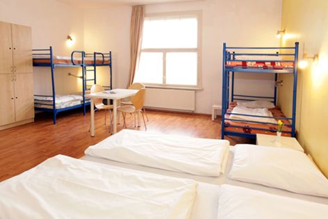 Blick auf ein Mehrbettzimmer in einem Hostel mit zwei blauen Etagenbetten, einem Doppelbett, einem Tisch und einem großen Fenster.