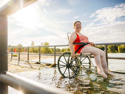 Bild von einer Rollstuhlfahrerin, die auf dem barrierefreien Badesteg am Cospudener See im Wasser steht und badet. 