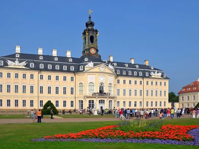 Wermsdorf - Schloss Hubertusburg, zu sehen ist eine Außenansicht des Schlosses, davor zahlreiche Menschen und eine mit roten und lila Blumen bepflanzte Grünfläche © Andreas Schmidt