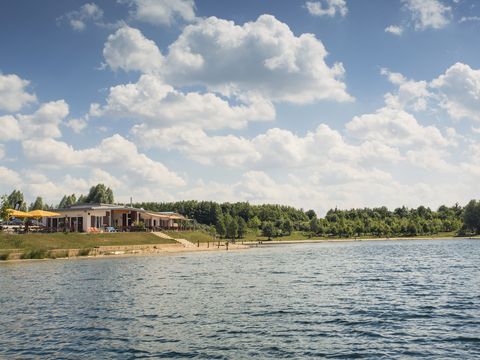 Blick auf das ALL-on-SEA Resort am Schladitzer See mit flachem, sandigen Ufer und umliegenden Grünflächen unter einem sonnigen Himmel mit vereinzelten Wolken.