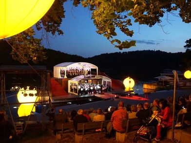 Seebühne Kriebstein, zu sehen sind ein Chor und ein Orchester, welche auf der Bühne musizieren sowie volle Zuschauerränge am Abend © Mittelsächsischer Kultursommer e.V.
